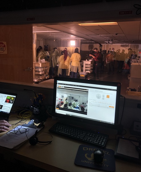 Computer monitor in the simulation lab control room showing action around a hospital bed with nurses in scrubs and gowns.